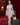 Realistic silicone doll in a white lace outfit standing in a room with red curtains and a lamp.