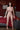 Sex doll standing in a bedroom setting with red curtains and a bed.