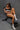 A highly realistic sex doll named Celeste, with long brown hair and a tan, is depicted sitting and holding a coffee cup. She is wearing a black and white sports crop top and leggings, exuding a sensual and health-focused aura.