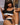 A highly realistic sex doll named Celeste, with long brown hair and a tan, is depicted sitting and holding a coffee cup. She is wearing a black and white sports crop top and leggings, exuding a sensual and health-focused aura.