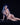 Naked person with blue hair and dark makeup sitting on a black surface against a black background