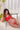 Person wearing a red lingerie set on a white couch with plants and curtains in the background.