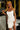 Woman in a white dress with braided hair standing in a bedroom setting.