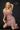 A highly realistic beige sex doll with long blonde hair, posed in a pink dress. The doll has a lifelike appearance with detailed facial features and a flexible skeleton, designed for versatile posing.
