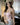 The image shows a highly realistic sex doll with long brown hair, wearing a pink lacey white top and a brown hat.