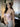 The image shows a highly realistic sex doll with long brown hair, wearing a pink lacey white top and a brown hat.