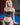 A realistic silicone doll with blonde hair, posed sitting next to a pool table, wearing a black bikini.