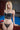 A realistic silicone doll with blonde hair, posed sitting next to a pool table, wearing a black bikini.