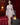 Realistic silicone doll in a white lace outfit standing in a room with red curtains and a lamp.