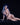 Naked person with blue hair and dark makeup sitting on a black surface against a black background