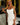 Woman in a white dress with braided hair standing in a bedroom setting.