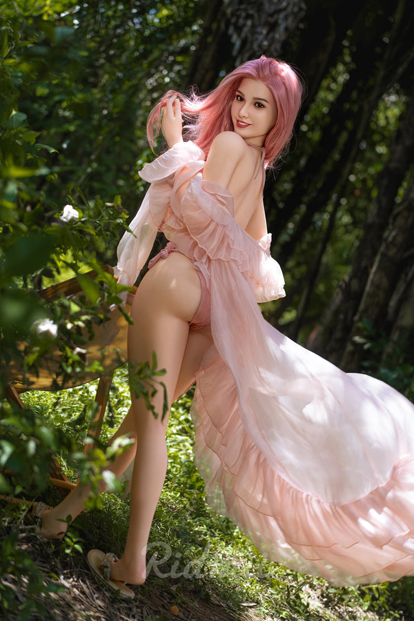 Woman in a flowing pink dress with pink hair standing in a natural setting.